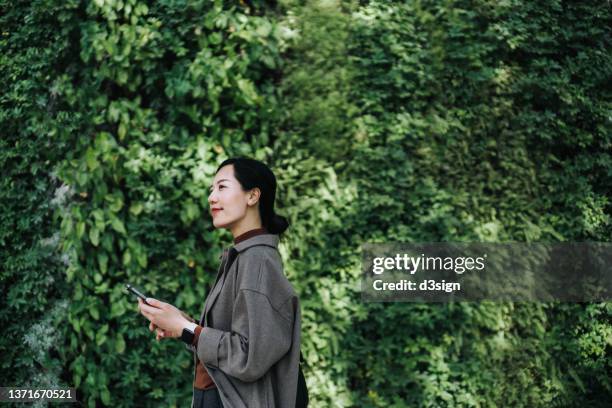 young asian woman looking the way forward while using smartphone outdoors, standing against wall covered with green plants and lush foliage. go green. green living, environmentally friendly lifestyle - public park design stock pictures, royalty-free photos & images