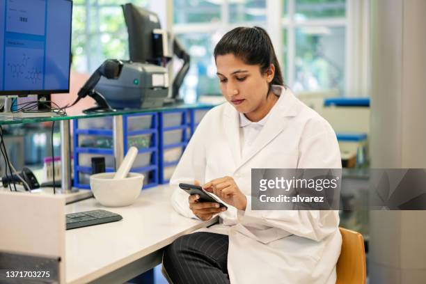asian woman scientist sitting at desk using her mobile phone - scientist standing next to table stock pictures, royalty-free photos & images