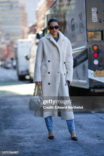 Guest wears brown sunglasses, a gray wool pullover, a white fluffy long coat, blue denim jeans pants, a white shiny leather puffy handbag from...