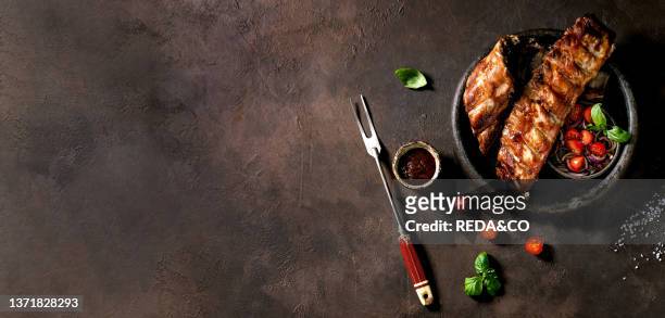 Grilled pork bbq ribs in ceramic plate served with meat fork. Marinated onions. Cherry tomatoes. Basil and barbeque sauce. Dark brown texture...