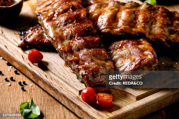 Grilled pork bbq ribs served with cherry tomatoes. Basil and barbeque sauce on wooden cutting board on wood board. Close up.