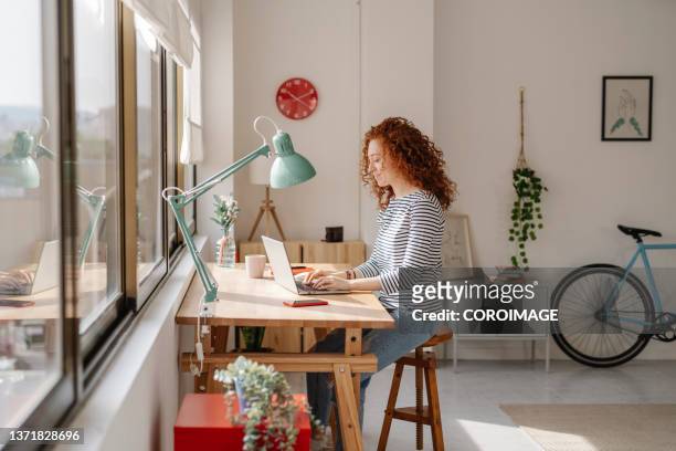 woman sitting on a desk using a laptop computer while working from home. - frau schreibtisch laptop büro stock-fotos und bilder