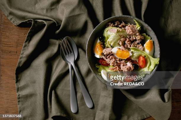 Traditional nicoise salad with canned tuna fish, olives and eggs, served in ceramic bowl with fork and spoon over wooden background with dark linen...