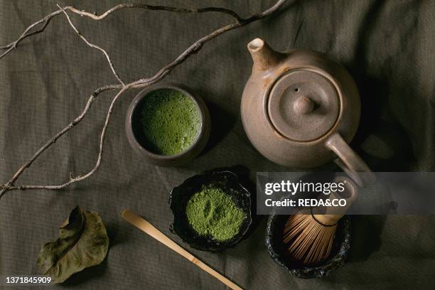 Traditional japanese hot green frothy tea matcha in ceramic cup, powdered matcha, teapot and bamboo whisk standing on dark linen tablecloth, Healthy...