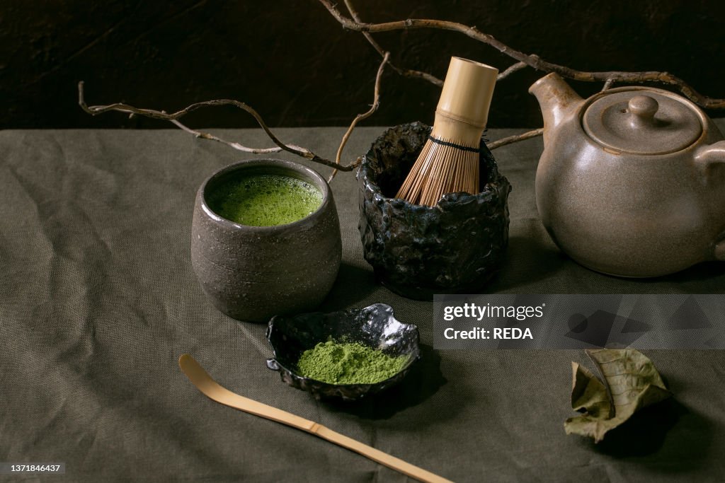 Traditional japanese hot green frothy tea matcha in craft ceramic cup, powdered matcha, teapot and bamboo whisk standing on dark linen tablecloth, Healthy drink