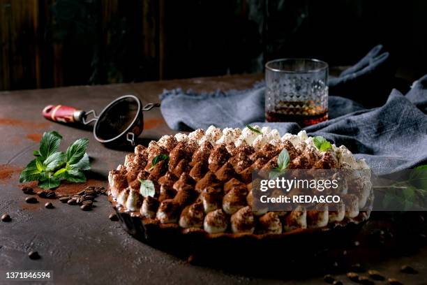 Homemade gluten free tiramisu traditional italian dessert sprinkled with cocoa powder decorated with mint leaves. Glass of whiskey. Blue textile...