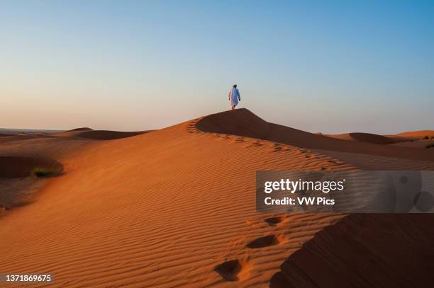 An Omani man walking along a sand dune ridge at sunset. Wahiba Sands, Arabian Peninsula, Oman..