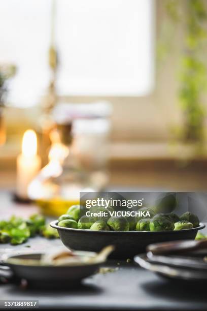 close up of green brussel sprouts in bowl at kitchen table with plates and blurred background with oil, candlelight and window with natural light - rosenkohl stock-fotos und bilder