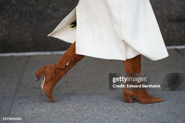 Guest wears a white long belted coat, brown suede pointed block heels knees boots / high boots, during New York Fashion Week, on February 14, 2022 in...