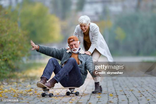 verspieltes seniorenpaar mit spaß auf dem skateboard im park. - candid mature couple outdoors stock-fotos und bilder