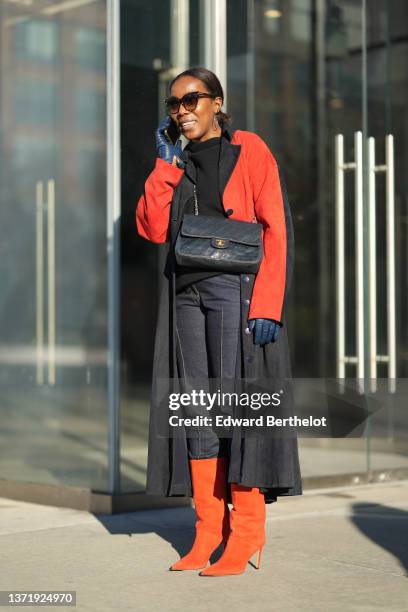 Guest wears sunglasses, gold earrings, a black high neck pullover, a bicolored red and black suede long coat, navy blue shiny leather gloves, a black...