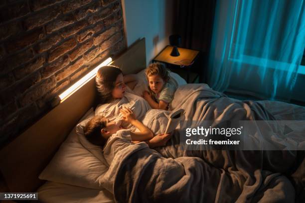 mother lying in bed and reading stories to her kids before sleep - kids sleep in bed stockfoto's en -beelden