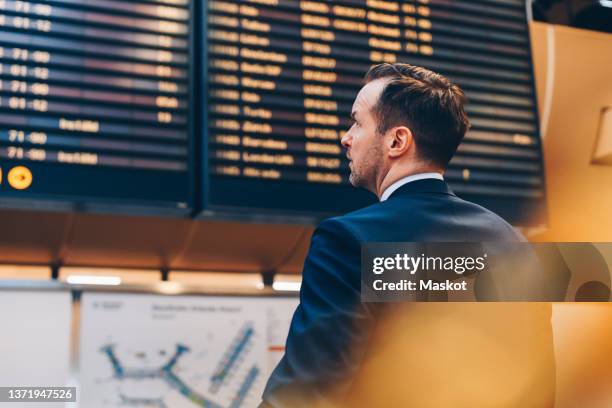 rear view of businessman looking at arrival departure board in airport - tabellone arrivi e partenze foto e immagini stock