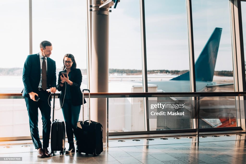Full length of business couple with luggage using mobile phone in airport