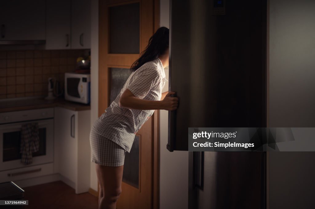 Woman leaning into fridge at night searching for food with bad diet habits.