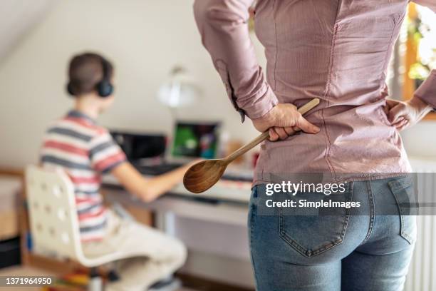 upset mother with hands on hips and wooden spoon in hand stands behind boy gaming on a computer with headphones on. - angry parent stock pictures, royalty-free photos & images