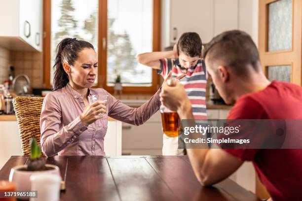 alcoholic couple drink and fight at the kitchen table while child standing in the background holds his ears. - family violence stock pictures, royalty-free photos & images