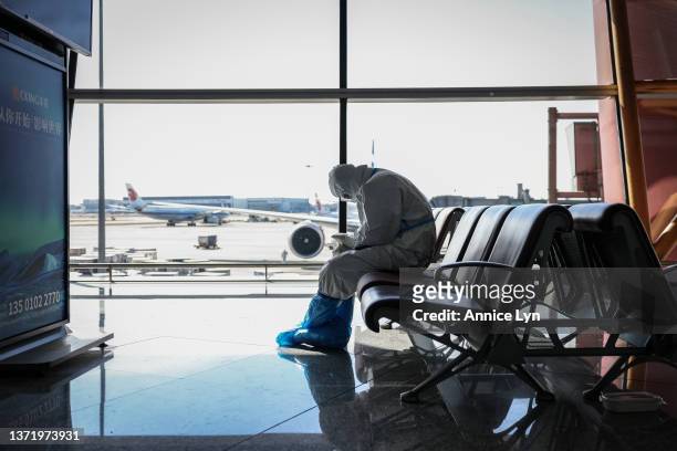 An airport staff member in a hazmat suit checks his phone at the Beijing Capital International Airport on February 21, 2022 in Beijing, China....