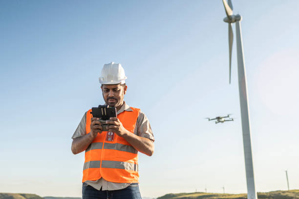 Man in a construction uniform piloting a drone by a wind turbine.