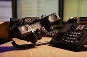 A man in black gloves with a telephone receiver in his hands, close-up. Office desk in a dark night room with computer monitors