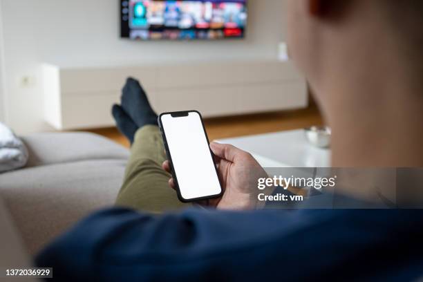 caucasian man holding a smart phone while watching tv - phone blank screen stock pictures, royalty-free photos & images