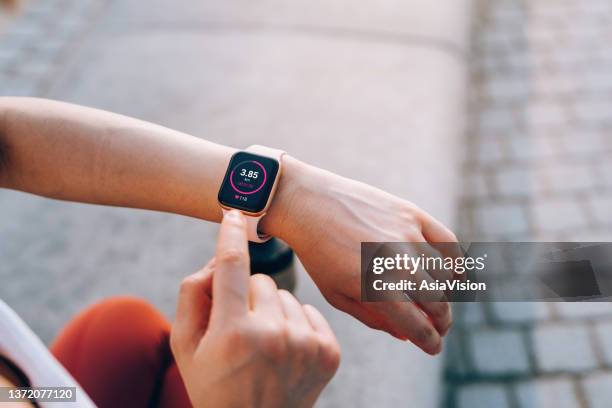 over the shoulder view of young asian sports woman checks her fitness statistics on smartwatch to monitor her training progress after working out in city outdoors. active and healthy lifestyle. outdoor workout. health and fitness with technology concept - taking pulse bildbanksfoton och bilder