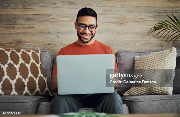 shot of a handsome young man sitting alone in his living room and using his laptop - man looking stock pictures, royalty-free photos & images