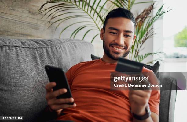 aufnahme eines gutaussehenden jungen mannes, der zu hause auf seinem sofa liegt und sein handy zum online-shopping benutzt - mann mit kreditkarte stock-fotos und bilder