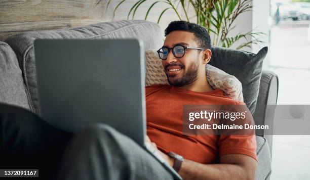 aufnahme eines gutaussehenden jungen mannes, der zu hause auf seinem sofa liegt und seinen laptop benutzt - zurücklehnen stock-fotos und bilder