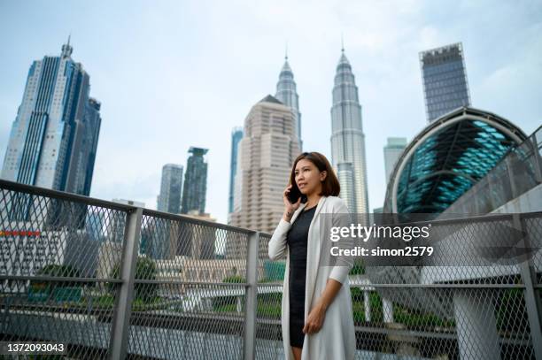 portrait of an asian woman talking on smartphone while standing in city bridge against city skyscrapers - malaysian people stock pictures, royalty-free photos & images