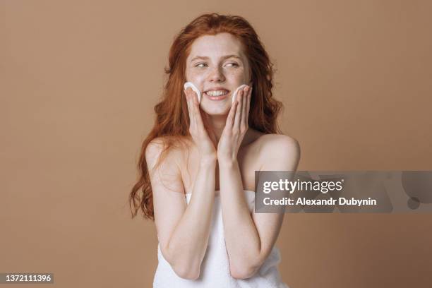 beautiful positive redhead woman with freckles takes care of her clean skin by rubbing with a cosmetic cotton pad. the concept of beauty and spa treatments. - facial cleanser stockfoto's en -beelden