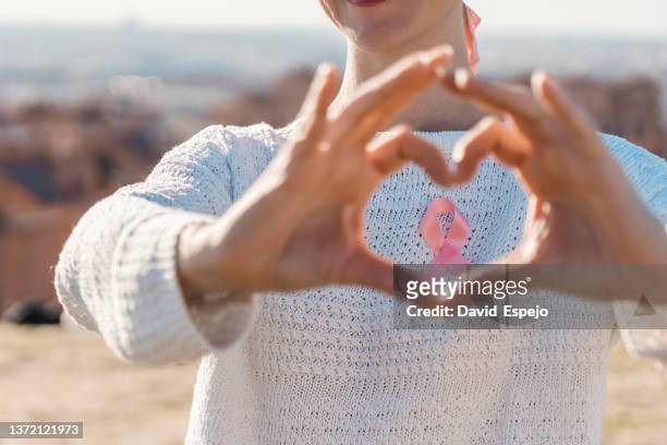 woman making a heart shaped with her hands while using a pink ribbon for support breast cancer cause. - fighting cancer stock pictures, royalty-free photos & images