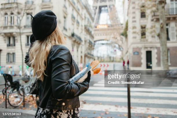 blonde frau, die an einem zebrastreifen steht, auf den eiffelturm in paris blickt, während sie zwei baguettes trägt - french baguette stock-fotos und bilder