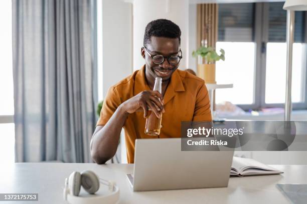 young man working from home on a laptop and drinking beer - man sipping beer smiling stockfoto's en -beelden