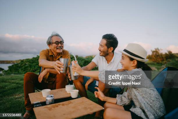 three generation family havinga a toast at campsite by the sea at dusk - asian smiling father son stock-fotos und bilder