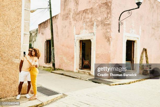 wide shot of smiling couple taking selfie on street corner while exploring town during vacation - couple standing stock pictures, royalty-free photos & images