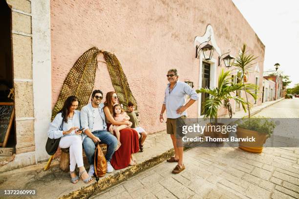 Wide shot of smiling multigenerational family relaxing on bench in front of boutique while exploring town during vacation