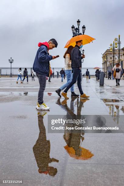 junger mann, der an regnerischen tagen auf den bildschirm seines mobiltelefons auf der plaza de la armeria schaut. um ihn herum laufen touristen mit sonnenschirmen und warmer kleidung über den platz - parasol de plage stock-fotos und bilder