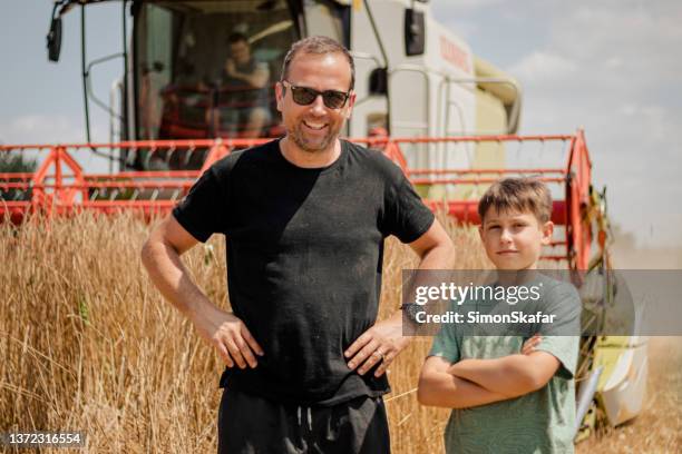 father and son standing next to wheat field in front of a combine harvester, smiling at camera - legacy stockfoto's en -beelden