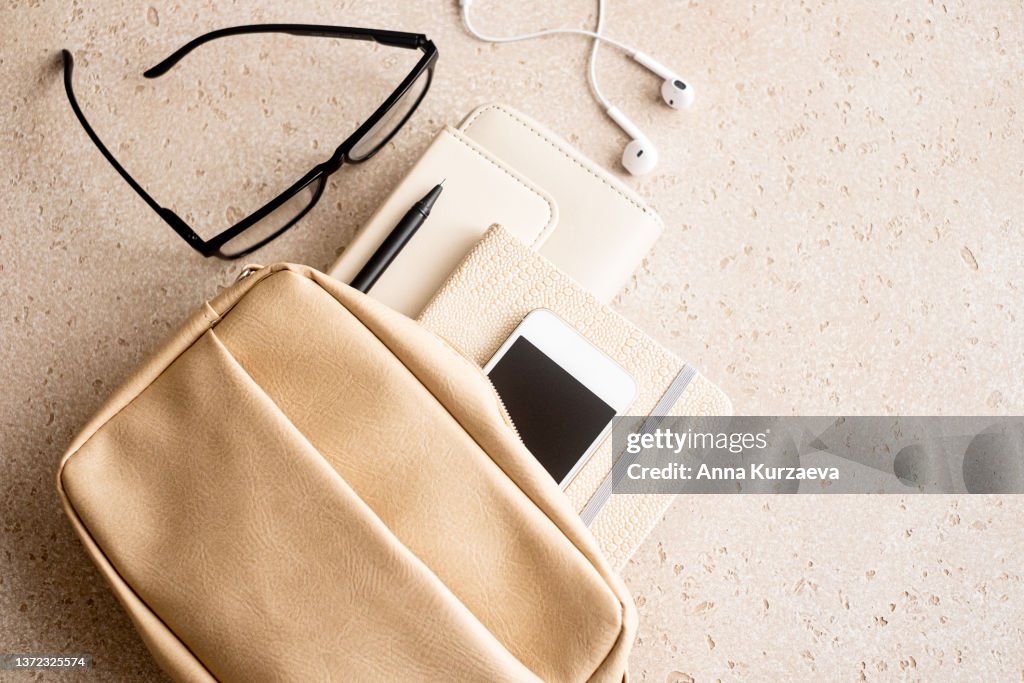 Beige woman handbag with purse, note pad, pen, smart phone, eyeglasses and in-ear headphones on concrete background from above, top view
