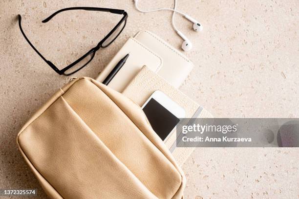 beige woman handbag with purse, note pad, pen, smart phone, eyeglasses and in-ear headphones on concrete background from above, top view - 手袋 個照片及圖片檔