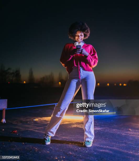 happy african young woman with headphones using mobile phone at night - one kid one world a night of 18 laughs stockfoto's en -beelden