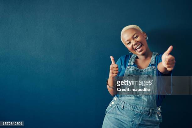 laughing woman wearing blue overalls giving the thumbs up on a blue background - excitement stock pictures, royalty-free photos & images