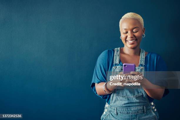 smiling woman in overalls texting on her phone against a blue background - short hair model stock pictures, royalty-free photos & images