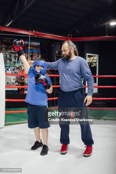 an armenian man with long hair stands in the boxing ring, holds the hand of his student who won the fight and looks at the camera - kids boxing stockfoto's en -beelden