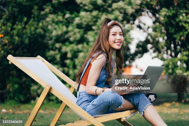 retrato de una hermosa y elegante joven asiática sosteniendo un teléfono inteligente, usando una computadora portátil mientras está sentada en una tumbona en un prado al aire libre en el parque. alegre joven mujer en mezclilla mirando a la cámara. dis - chinese female university student portrait fotografías e imágenes de stock