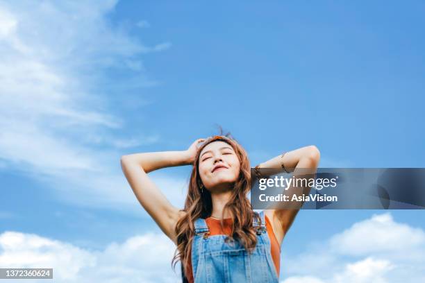 schöne junge asiatin mit geschlossenen augen und tiefem atemzug im freien in der natur, gegen blauen himmel an einem sonnigen tag. sonne und freiheit in der natur genießen - atmungsaktiv stock-fotos und bilder