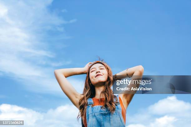 beautiful young asian woman with eyes closed open arms and taking deep breath outdoors in nature, against blue sky on a sunny day. enjoying sunshine and freedom in nature - air escaping stock pictures, royalty-free photos & images