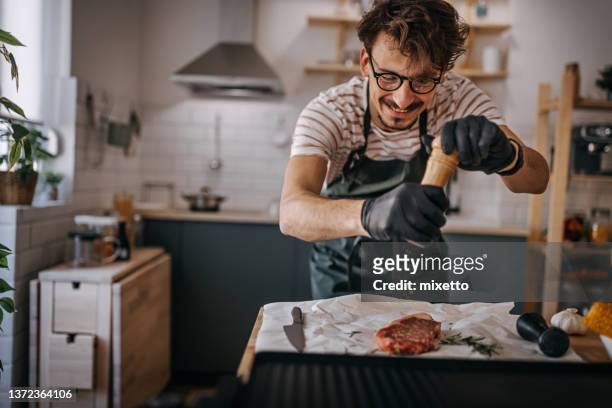 young man sprinkling pepper on steak in kitchen - strooisels stockfoto's en -beelden
