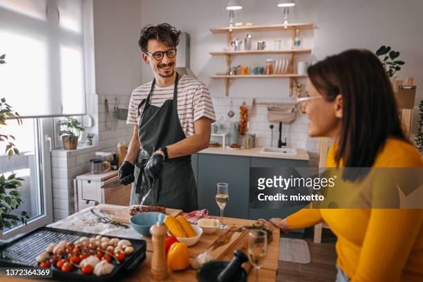 uomo sorridente che parla con la ragazza mentre cucina il cibo in cucina - couple in kitchen foto e immagini stock